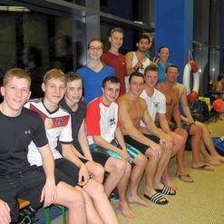 Gruppe von Jugendlichen in Badekleidung, sitzend auf einer Bank in einem Schwimmbad, lächelnd in die Kamera.