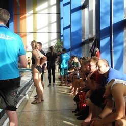 Gruppe von Schwimmern in Badekleidung sitzt am Beckenrand eines Schwimmbads, während Trainer Anweisungen gibt.