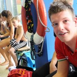 Junger Mann in rotem Shirt sitzt neben Schwimmern auf einer Bank in einem Schwimmbad, Rettungsring im Hintergrund.