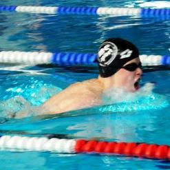 Junger Schwimmer mit Badekappe und Sonnenbrille sprintet in einem Wettkampfbecken. Spritzwasser um ihn herum.