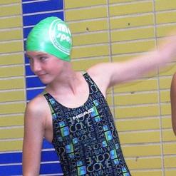 Junge Schwimmerin mit grünem Badekappe und Badeanzug, steht vor einer farbigen Fliesenwand und gestikuliert.