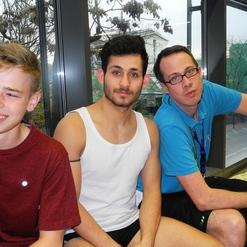 Drei junge Männer sitzen in einem Raum mit großen Fenstern, lächelnd und entspannt nebeneinander.