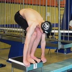 Schwimmer in schwarzer Badehose und Badekappe springt vom Startblock ins Wasser.