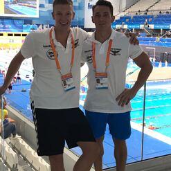Zwei Männer in Stadionkleidung stehen am Rand eines Schwimmbeckens und lächeln in die Kamera.