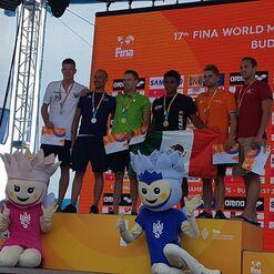 Sieben Medaillengewinner stehen auf einer Bühne bei den FINA World Masters in Budapest, umgeben von Maskottchen und Bannern.
