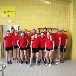 Gruppe von 15 Personen in roten T-Shirts vor einer gelben Fliesenwand im Schwimmbadbereich.