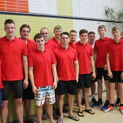 Gruppe von zwölf Personen in roten T-Shirts, stehend vor einer hellen Wand in einem Schwimmbeckenbereich.