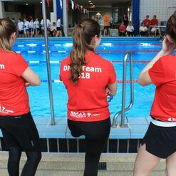Drei Frauen in roten T-Shirts mit der Aufschrift „DMS-Team 2018“ stehen am Beckenrand eines Schwimmbads.