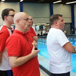 Vier Schwimmtrainer in roten T-Shirts stehen am Beckenrand und feuern Schwimmer an. Das Schwimmbecken ist im Hintergrund sichtbar.