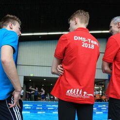 Drei Personen stehen vor einem Schwimmbecken, tragen Team-Shirts in Blau und Rot mit "DMS-Team 2018".