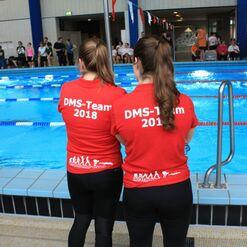 Zwei Personen mit roten T-Shirts stehen am Schwimmbecken und beobachten den Wettkampf. Auf den T-Shirts steht „DMS-Team 2018“.