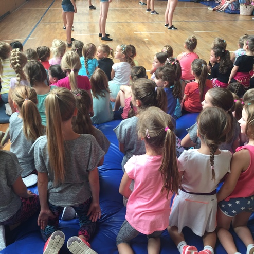 Mädchen in bunten Shirts sitzen auf einer blauen Matte und schauen einem Auftritt in einer Sporthalle zu.