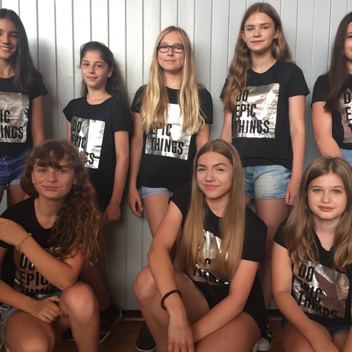 Gruppenfoto von acht Mädchen in schwarzen T-Shirts mit dem Aufdruck "DO EPIC THINGS", lächelnd vor einer hellen Wand.