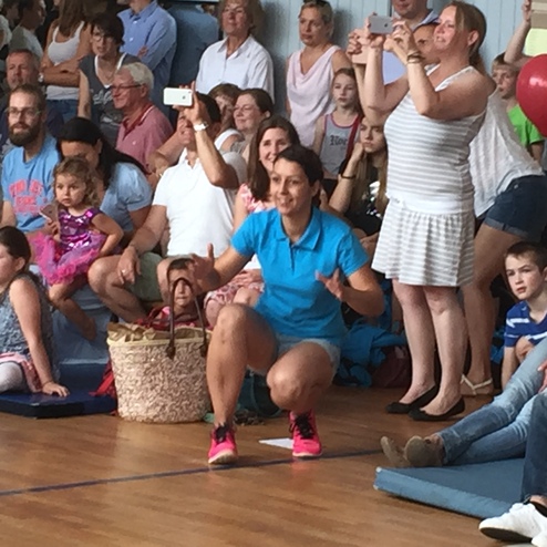 Eine Gruppe von Menschen sitzt und steht in einer Halle, einige filmen oder fotografieren eine Sportvorführung.