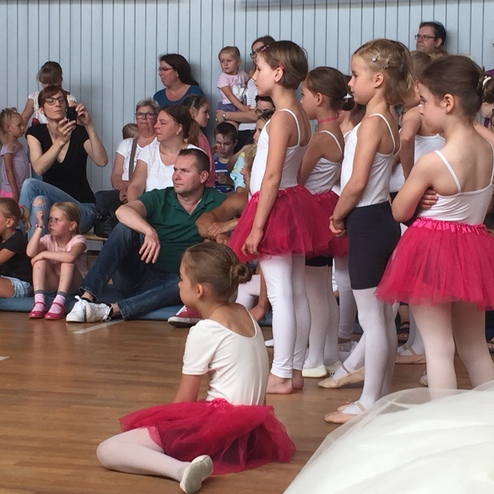 Gruppe junger Tänzerinnen in Ballettanzug und Tüllröcken bei einer Aufführung, umgeben von Zuschauern.