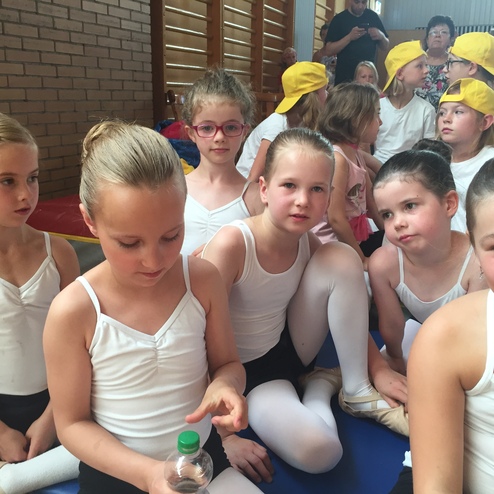 Gruppe von Mädchen in Ballettkleidung, einige halten Wasserflaschen, sitzen auf einer Matte in einer Turnhalle.
