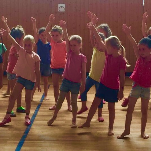 Gruppe von Mädchen in bunten T-Shirts und Shorts, die im Tanzstudio synchron tanzen und dabei die Arme heben.
