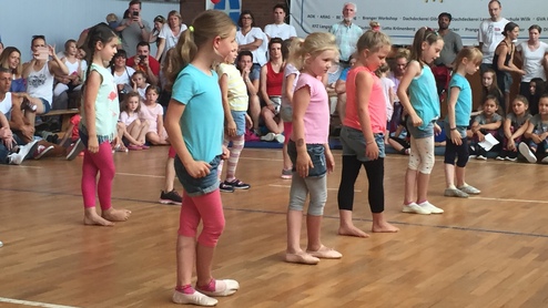 Sechs Mädchen tanzen auf einer Bühne, vor einer Zuschauermenge mit Farbenfrohen Ballons im Hintergrund.