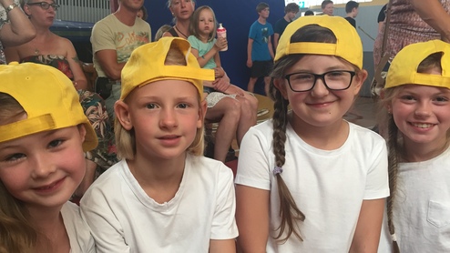 Vier Kinder mit gelben Caps lächeln in weißen T-Shirts, während Erwachsene im Hintergrund zuschauen.