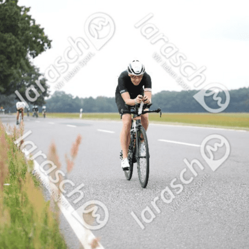 Triathletin in schwarzer Radkleidung fährt konzentriert auf der Straße, umgeben von grünem Gras und weiteren Radfahrern.