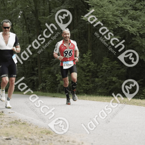 Zwei Männer joggen auf einem Waldweg, einer im schwarzen, der andere im roten Trikot, umgeben von Bäumen.