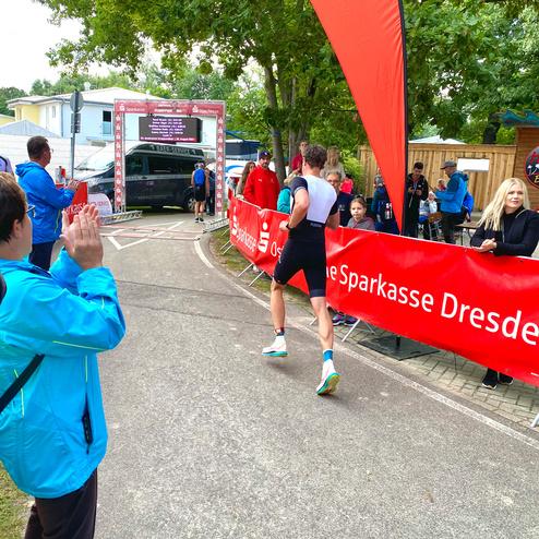 Läufer überquert die Ziellinie einer Veranstaltung, umgeben von jubelnden Zuschauern und Werbebannern.