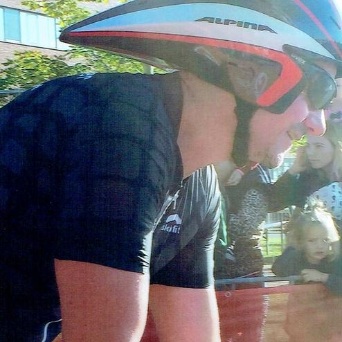 Ein Radfahrer in einem aerodynamischen Helm fährt vorbei, während Zuschauer hinter einer roten Absperrung zuschauen.