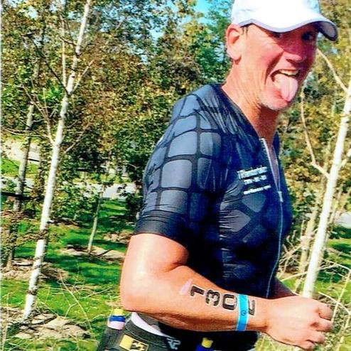 Läufer trägt sportliche Bekleidung und startet auf einem Weg mit Bäumen im Hintergrund. Er zeigt die Zunge.