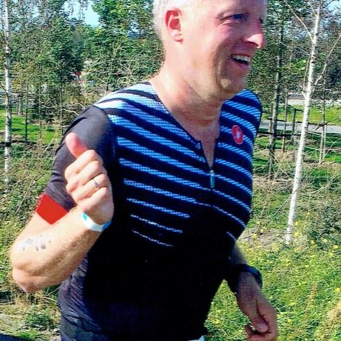 Läufer in sportlicher Kleidung mit Nummernband joggt auf einem Weg, umgeben von Bäumen und Wiesen.