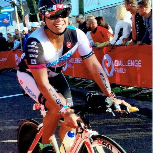 Sportler auf einem Zeitfahrrad in einem Wettkampf, mit einem Wasserbehälter und Zuschauern im Hintergrund.