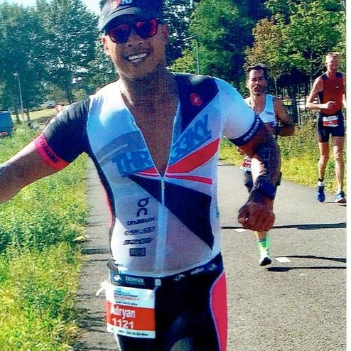 Läufer in sportlicher Kleidung mit Startnummer beim Triathlon, grinsend auf einer Strecke umgeben von Bäumen.