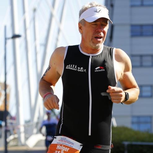 Mann in Wettkampfanzug läuft an einer Uferpromenade, trägt eine Startnummer und einen weißen Sonnenschutz.