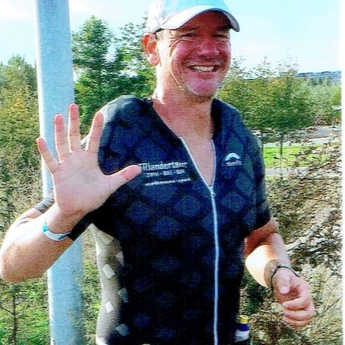 Mann in Sportbekleidung winkt lächelnd beim Laufen auf einem Weg entlang von Bäumen und Gras.