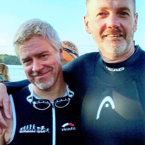 Zwei Männer in Sportanzügen stehen nebeneinander am Seeufer, lächeln und posieren für ein Foto.