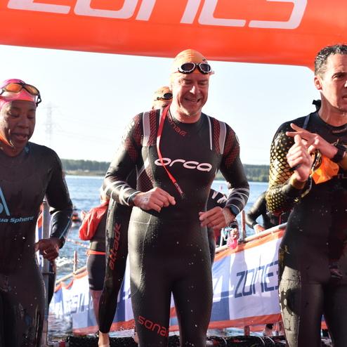 Drei Schwimmer in Neoprenanzügen stehen am Ufer, hinter ihnen ist ein orangefarbener Zielbogen sichtbar.