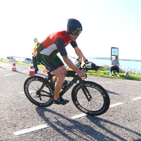Athlet in Radrennsportkleidung fährt auf einem schwarzen Rennrad entlang einer Straße mit Zuschauern im Hintergrund.
