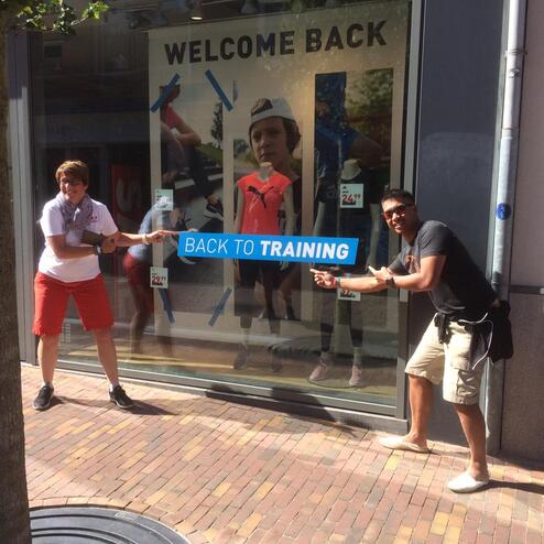 Zwei Personen zeigen lächelnd auf ein Werbeschild "BACK TO TRAINING" in einem Schaufenster.