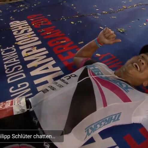 Mann liegt erschöpft auf dem Boden einer Sportveranstaltung, umgeben von Konfetti und Werbebannern.