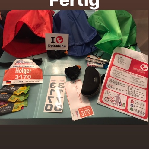 Sammlung von Triathlon-Ausrüstung: Startnummer, Gels, Flyer und bunte Taschen auf einem Tisch.