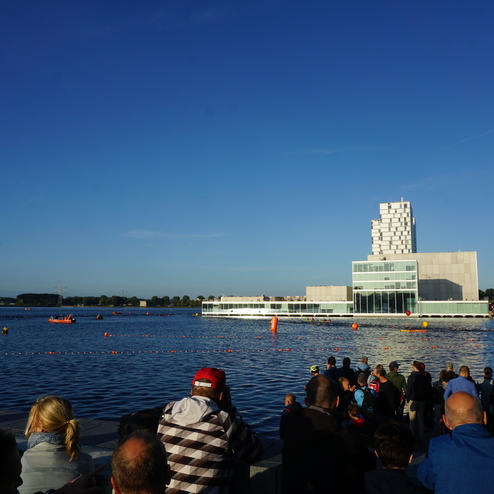 Menschen beobachten ein Wasserrennen mit Booten auf einem Gewässer vor einem modernen Gebäude bei klarem Himmel.