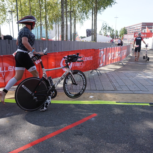Teilnehmer beim Triathlon schiebt sein Rennrad auf einem markierten Weg, umgeben von Zuschauerständen und Werbebannern.