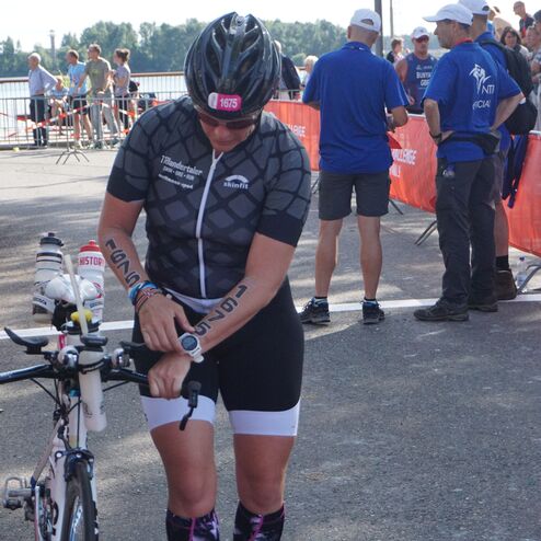 Sportlerin in Wettkampfkleidung justiert ihre Uhr an einem Rennrad, im Hintergrund Zuschauer und Event-Szenen.