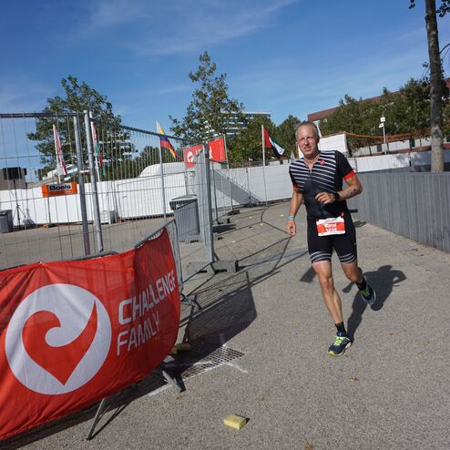 Athlet in sportlicher Kleidung läuft an einem roten Banner der Challenge Family vorbei, umgeben von Bäumen und Absperrungen.