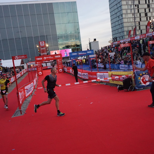 Läufer überquert die Ziellinie bei einem Triathlon, umgeben von Zuschauern auf einem roten Teppich.