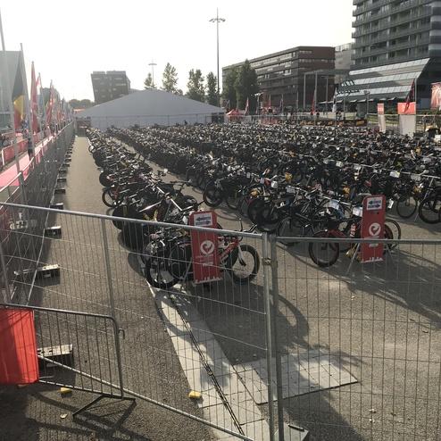 Große Fahrradabstellfläche mit vielen Fahrrädern, umgeben von Absperrungen und Fahnen, unter klarem Himmel.