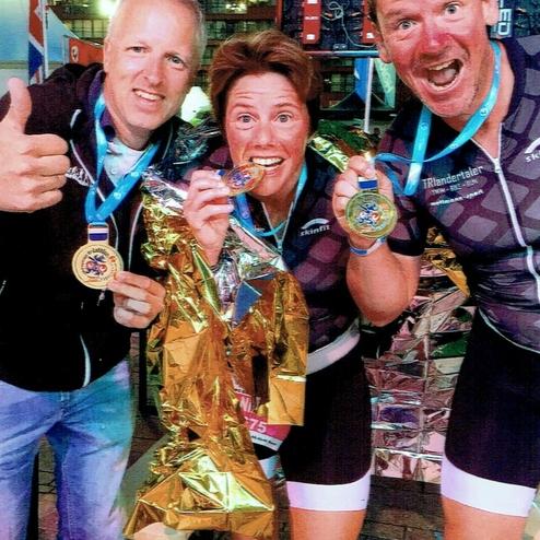 Drei Personen zeigen begeistert ihre Medaillen vor einer bunten, metallischen Kulisse nach einem Wettkampf.