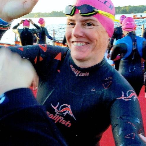 Lächelnde Person in einem Neoprenanzug, mit rosa Badekappe, an einem Wasserstartpunkt mit Schwimmern im Hintergrund.
