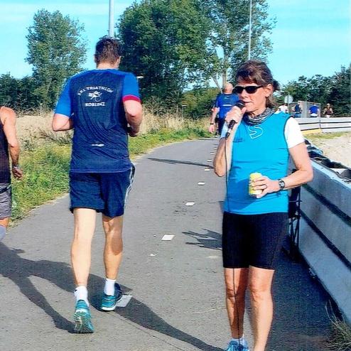 Frau mit Mikrofon auf einer Laufstrecke, im Hintergrund laufende Teilnehmer und grüne Landschaft.