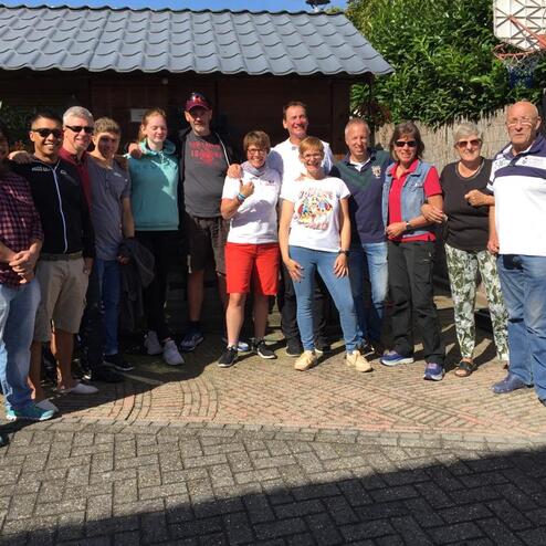 Gruppe von 15 Personen posiert vor einem Gartenhaus, umgeben von Pflanzen und einem Basketballkorb.