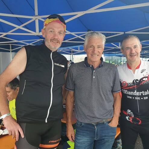 Drei Radfahrer stehen in einer Sportveranstaltung unter einem blauen Zelt und lächeln in die Kamera.